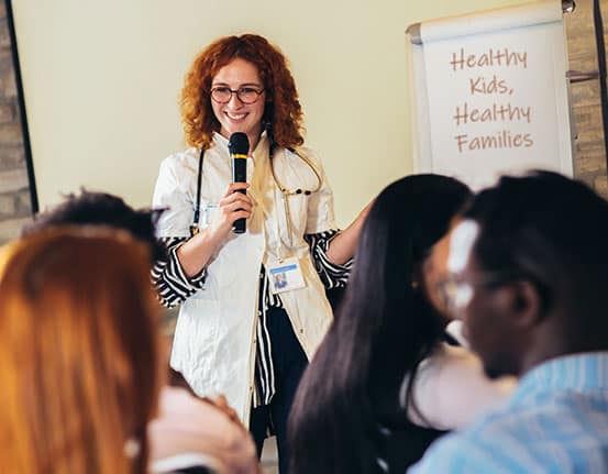 A doctor stands in front of a crowd with a microphone in hand