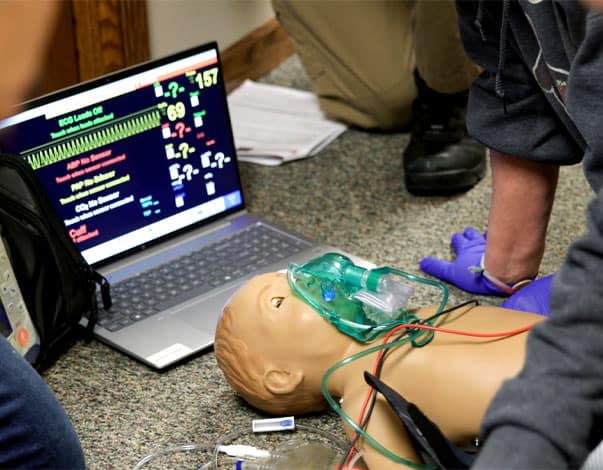 A pediatric manikin ls on a carpeted floor with breathing equipment on its face and a computer displaying metrics in the background